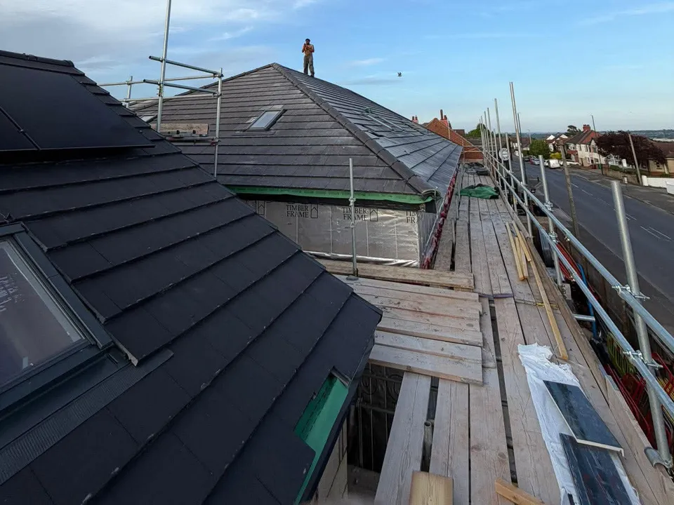 A roofer at work on a dark tiled roof under a blue sky